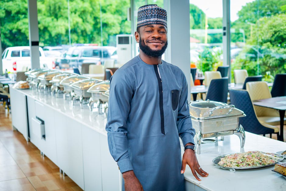 African chef in traditional attire smiling in a bright restaurant with buffet setup.
